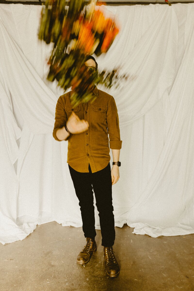 Anthony Gauna stands against a white cloth backdrop in a mustard shirt and boots as a bouquet of flowers flies past his face