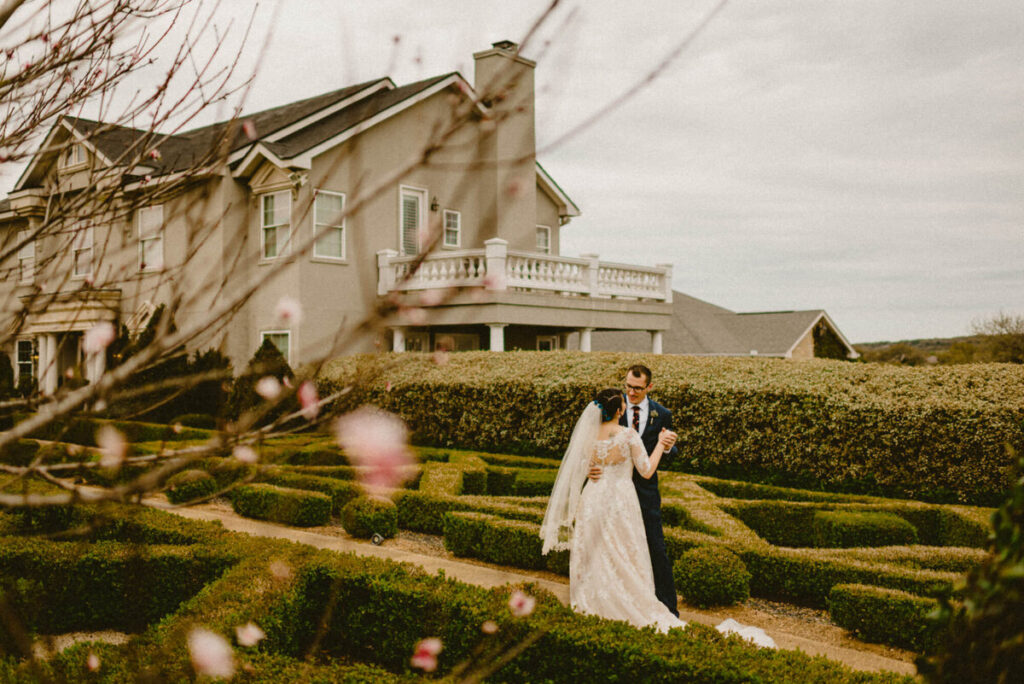 A bride in a lace gown and a groom in a navy suit walk through a boxwood garden maze in front of their wedding venue, seen through blossoming branches